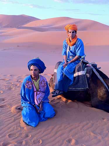 en la imagen son dos niños bereberes uno sentado sobre la arena del desierto y el otro sobre un camello estan vestidos de color azul